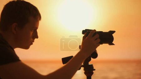 Dieses Bild zeigt die Silhouette eines männlichen Fotografen, der in der goldenen Stunde in das Fotografieren vertieft ist. Der warme Sonnenuntergang im Hintergrund unterstreicht den friedlichen Moment der Kreativität.