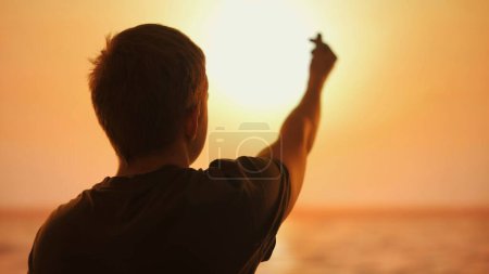 Silhouette of a man on sunset background making Korean heart sign with fingers raising his hand to the sunset sun, symbolizes love and connection with nature.