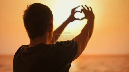 This image features a mans silhouette against a sunset, forming a heart shape with his hands around the sun, symbolizing love and connection with nature.