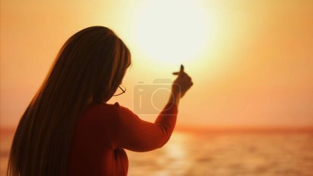 Silhouette of a woman on sunset background making Korean heart sign with fingers raising her hand to the sunset sun, symbolizes love and connection with nature.