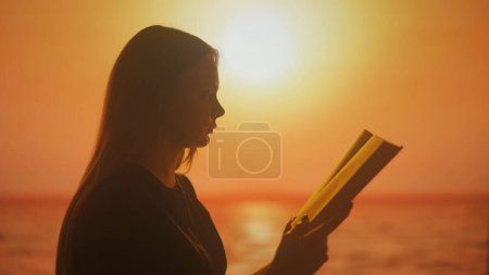 Foto de Una silueta de una joven mujer leyendo un libro sobre un fondo de brillantes tonos de puesta de sol. La imagen transmite la esencia de la inspiración extraída de la belleza de la naturaleza - Imagen libre de derechos
