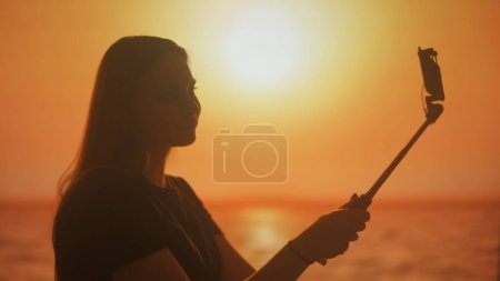 Foto de Silueta de una joven mujer hablando en una videollamada en un teléfono inteligente en el fondo de tonos brillantes de la puesta del sol. Comunicación en línea, transmisión en vivo en las redes sociales. - Imagen libre de derechos