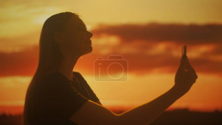 Silhouette of a young female taking selfies with a smartphone against the bright colors of the sunset. The tranquil scene evokes a sense of peace and lightness.