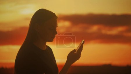 Silhouette einer jungen Frau, die Informationen auf einem Smartphone vor den grellen Farben des Sonnenuntergangs durchsucht. Die ruhige Szenerie vermittelt ein Gefühl von Frieden und Leichtigkeit.