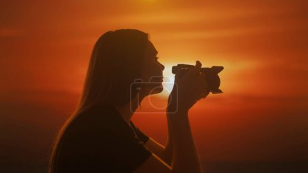 Foto de Silueta de una joven hembra tomando una foto con una cámara sobre el fondo de brillantes tonos de atardecer. - Imagen libre de derechos