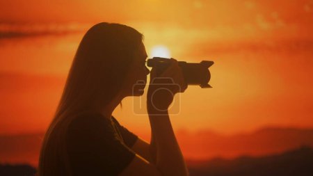 Téléchargez les photos : Silhouette d'une jeune femelle prenant une photo avec un appareil photo sur fond de nuances lumineuses de coucher de soleil. - en image libre de droit