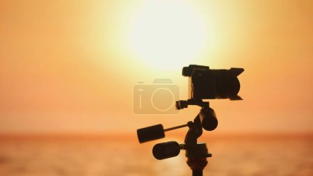 Die markante Silhouette einer professionellen Kamera auf einem Stativ fängt die Essenz des Filmemachens vor dem Hintergrund eines lebendigen Sonnenuntergangs ein. Kunst, Momente festzuhalten.