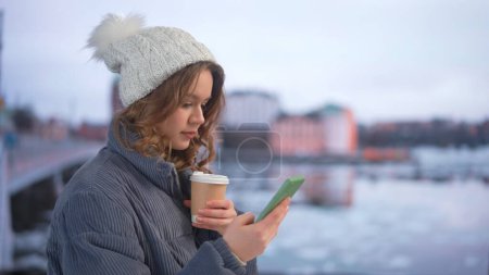 Photo for Gadgets and modern everyday life advertisement concept. Female using smartphone on daily basis. Young woman in knit hat standing on the street drinking coffee and watching social media online. - Royalty Free Image