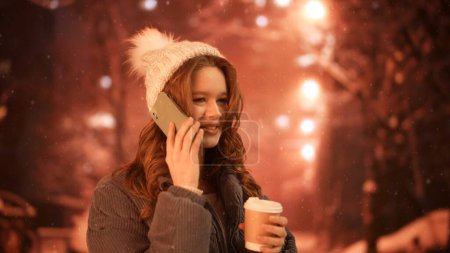 Photo for Gadgets and modern everyday life advertisement concept. Female using smartphone on daily basis. Young woman in knit hat on the street at night drinking coffee and talking with friend on smartphone. - Royalty Free Image