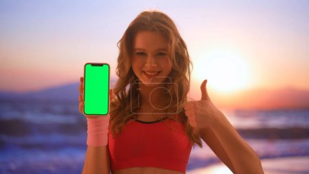 Gadgets und moderne Werbekonzepte für den Alltag. Frau mit Smartphone auf täglicher Basis. Junge Frau in Sportbekleidung am Strand bei Sonnenuntergang zeigt auf Smartphone-Bildschirm.