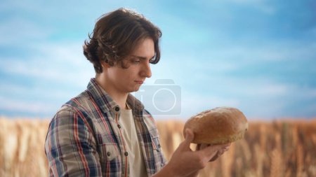 Photo for Agriculture and modern technology concept. Farmer against large field of wheat at sunset. Man agronomist standing in the farm field, holding in hands and giving fresh loaf of bread. - Royalty Free Image