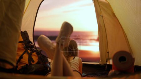 Téléchargez les photos : Concept camping et aventure. Personne en camping voyage et randonnée, explorer la nature. Jeune femme couchée à l'intérieur de la tente au coucher du soleil sur la plage regardant à l'extérieur, vacances et vacances en plein air. - en image libre de droit