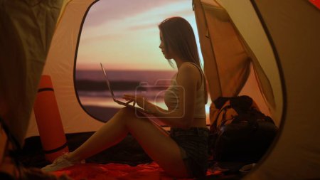 Photo for Camping and adventure concept. Person on campsite traveling and hiking, exploring nature. Young woman sitting inside the tent at sunset on the beach working on laptop, holiday and vacation outdoors. - Royalty Free Image