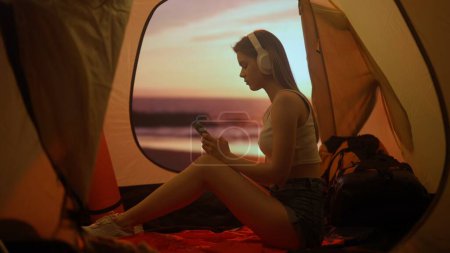 Téléchargez les photos : Concept camping et aventure. Personne en camping voyage et randonnée, explorer la nature. Jeune femme assise dans un casque écoutant de la musique sur smartphone à l'intérieur de la tente au coucher du soleil sur la plage. - en image libre de droit
