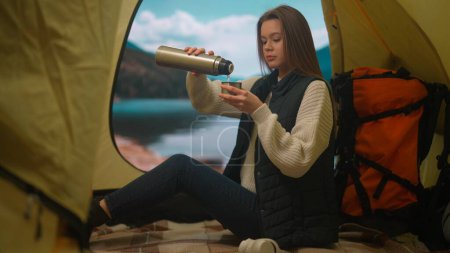 Téléchargez les photos : Personne dans un camping voyage et randonnées, explore la nature. Une jeune femme s'assoit et se verse le thé d'un thermos dans une tente au bord d'un lac dans les montagnes, se relaxant dans la nature - en image libre de droit
