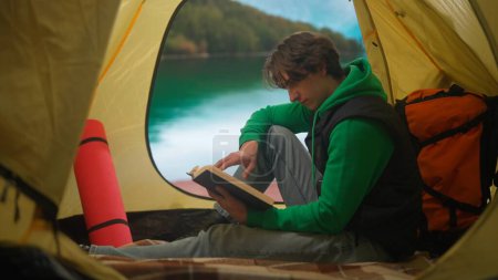 Téléchargez les photos : Concept camping et aventure. Personne en camping voyage et randonnée, explorer la nature. Jeune homme assis et lire le livre à l'intérieur de la tente au bord du lac dans les montagnes, vacances en plein air. - en image libre de droit