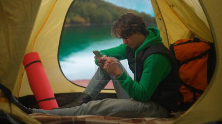 Foto de El concepto de camping y aventura. Una persona en un camping viaja y caminatas, explora la naturaleza. Un joven está enviando mensajes en su teléfono inteligente en una tienda de campaña en la orilla de un lago en las montañas. - Imagen libre de derechos
