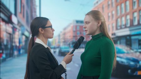 Photo for Live tv news broadcasting concept. Female presenter reporting on air outside. Woman news host journalist taking interview with young female on the street, city background at the back. - Royalty Free Image