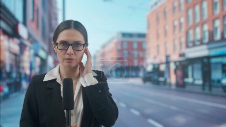 Photo for Live tv news broadcasting concept. Female presenter reporting on air outside. Woman news host journalist talking with studio, wearing earphone, standing on the street, city background at the back. - Royalty Free Image