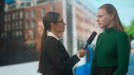 Photo for Live tv news broadcasting concept. Female presenter reporting on air outside. Woman news host journalist taking interview with young female on the street, city background at the back. - Royalty Free Image