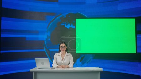 Téléchargez les photos : Live tv news concept de diffusion. Présentatrice dans le studio. Femme hôte de nouvelles est assis au bureau présentant des événements quotidiens, reportage sur l'air, chroma écran vert clé à l'arrière. - en image libre de droit