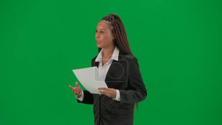 Photo for Tv news report and live broadcasting advertisement concept. Female reporter isolated on chroma key green screen background. African American woman news host in suit reads paper documents and talks - Royalty Free Image