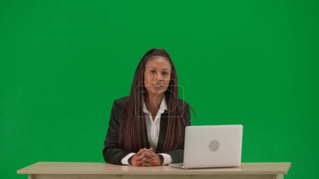 Photo for Tv news report and live broadcasting advertisement concept. Female reporter isolated on chroma key green screen background. African American woman news host in suit at the desk with laptop. - Royalty Free Image