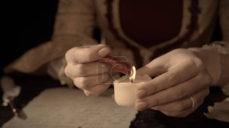 Historical letter creative concept. Female in antique outfit at the desk with candle. Woman in renaissance dress holding wax stick and warms it on a candle flame.