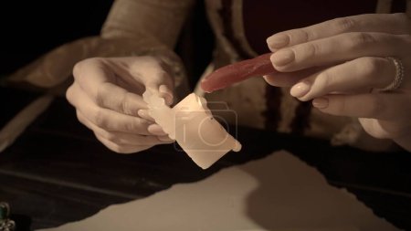 Historical letter creative concept. Female in antique outfit at the desk with candle. Close up shot of woman holding wax stick and warms it on a candle flame
