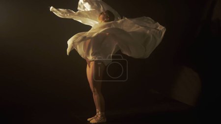 Photo for Essence of dance through the delicate interplay of light and shadow, as a dancer shrouded in white chiffon moves with poise. The subtle lighting underscores the fluidity and grace of dancers motion - Royalty Free Image