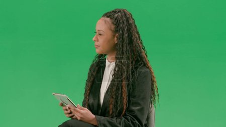 Tv news and live broadcasting concept. Young female reporter isolated on chroma key green screen background. African american woman tv news host sit in chair with tablet taking interview.