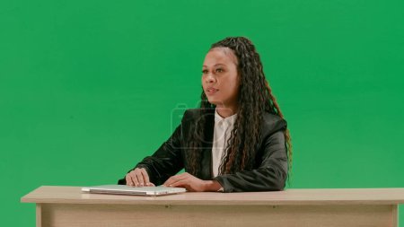 Tv news and live broadcasting concept. Female reporter at the desk isolated on chroma key green screen background. African american woman tv news host sitting talking at camera.