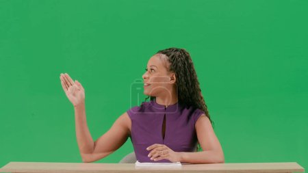 Tv news and live broadcasting concept. Female in dress sit at the desk isolated on chroma key green screen background. African american woman tv news host sitting talking looking at camera.