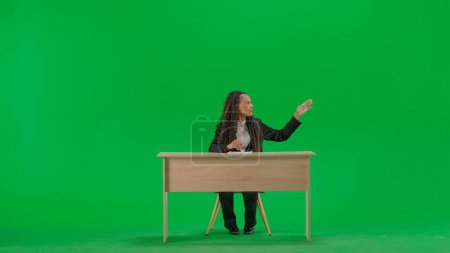 Photo for Tv news and live broadcasting concept. Female reporter at the desk isolated on chroma key green screen background. Full shot african american woman tv news host sitting talking at camera. - Royalty Free Image