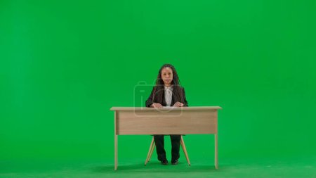 Tv news and live broadcasting concept. Female reporter at the desk isolated on chroma key green screen background. Full shot african american woman tv news host sitting talking at camera.