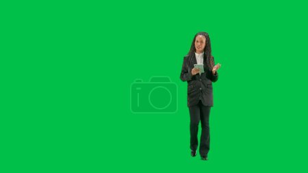Photo for Tv news and live broadcasting concept. Young female reporter isolated on chroma key green screen background. Full shot african american woman tv news host walking holding tablet and talking. - Royalty Free Image