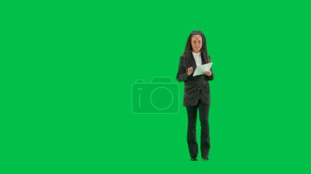TV-Nachrichten und Live-Übertragungskonzept. Junge Reporterin isoliert auf grünem Hintergrund. Full shot afrikanisch-amerikanische Frau tv news host walking talking and reading papers.