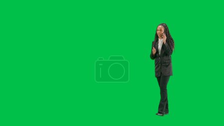 Téléchargez les photos : Nouvelles TV et concept de diffusion en direct. Jeune reporter femelle isolée sur fond vert chromatique. Full shot afro-américaine femme tv nouvelles hôte marche et parler sur smartphone. Demi-tour. - en image libre de droit