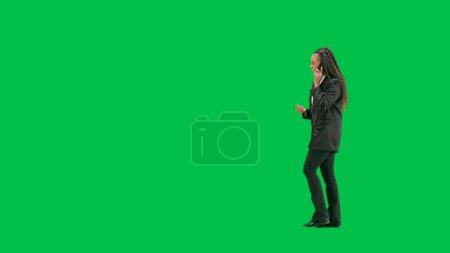 Photo for Tv news and live broadcasting concept. Young female reporter isolated on chroma key green screen background. Full shot african american woman tv news host walking and talking on smartphone. Side view. - Royalty Free Image