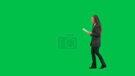 Photo for Tv news and live broadcasting concept. Young female reporter isolated on chroma key green screen background. Full shot african american woman tv news host walking talking and reading papers. Side view - Royalty Free Image