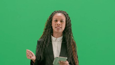 Tv news and live broadcasting concept. Young female reporter isolated on chroma key green screen background. African american woman tv news host walking holding tablet and talking.
