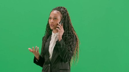 Photo for Tv news and live broadcasting concept. Young female reporter isolated on chroma key green screen background. African american woman tv news host walking and talking on smartphone. Half turn. - Royalty Free Image