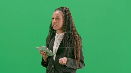 Photo for Tv news and live broadcasting concept. Young female reporter isolated on chroma key green screen background. African american woman tv news host walking holding tablet and talking. Half turn. - Royalty Free Image