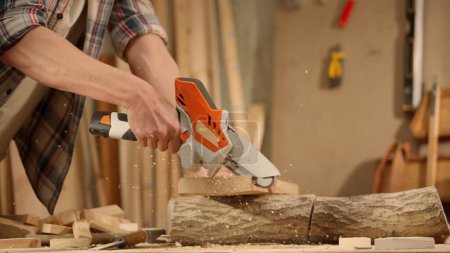 Photo for Carpentry and handicraft advertisement concept. Male woodworker working in garage. Man professional carpenter working with wooden materials in workshop sawing wood using cordless mini chainsaw. - Royalty Free Image