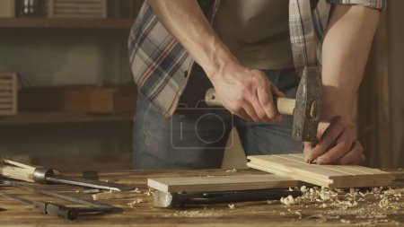 Foto de Concepto de publicidad de carpintería y artesanía. Hombre carpintero trabajando en garaje. Hombre carpintero profesional trabajando con materiales de madera en taller y golpeando clavos en panel de madera con mazo. - Imagen libre de derechos