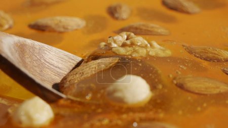 Photo for Food advertisement concept. Healthy organic honey. Sweet fresh golden honey on the yellow background, wooden spoon scooping almonds walnuts and cashew nuts covered in the thick syrup, close up shot. - Royalty Free Image