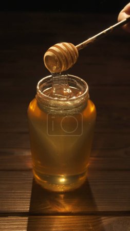 Téléchargez les photos : Concept de publicité alimentaire. Miel bio sain dans un pot. Pot en verre transparent avec doux miel doré scintillant au soleil, trempette au miel à l'intérieur du pot sur la table en bois, plan complet. - en image libre de droit