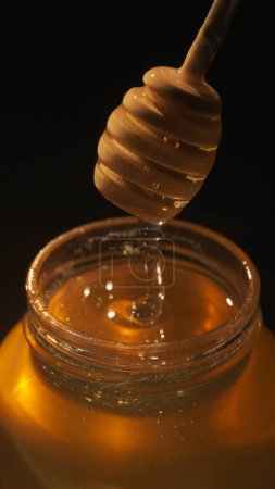 Téléchargez les photos : Concept de publicité alimentaire. Miel bio sain dans un pot. Pot en verre transparent avec doux miel doré scintillant au soleil, trempette au miel à l'intérieur du pot sur la table en bois, fermer. - en image libre de droit