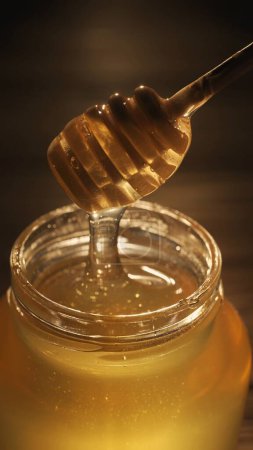 Photo for Food advertisement concept. Healthy organic honey in a jar. Transparent glass jar with sweet golden honey sparkling in the sun, honey dipper inside jar on the wooden table, close up. - Royalty Free Image