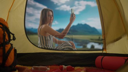 Téléchargez les photos : Concept camping et aventure. Femme reposant sur le camping relaxant près du lac. Jeune femme assise devant la tente à la lumière du jour dans la côte du lac de montagnes, parlant par appel vidéo sur smartphone, - en image libre de droit